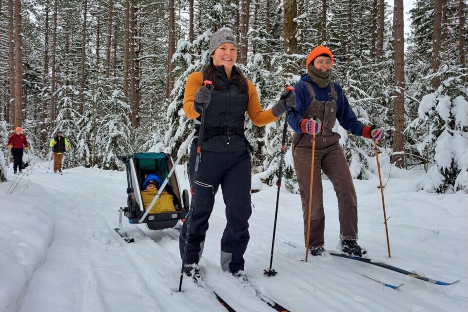 Two people nordic skiing along the All Persons Trail. One pulls a sled with a baby.