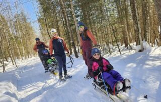 A group of smiling people stand on a snowy trail. Three are adults in snowshoes and matching jackets, two are smiling children in colorful clothing sitting in sit-ski assistive devices.